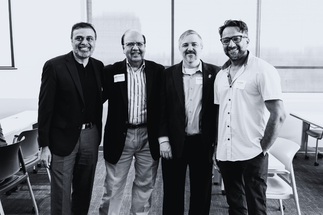 Four men pose together for a photo in a bright conference room. They are smiling and dressed in business attire, with one man wearing glasses, indicating a professional networking event.